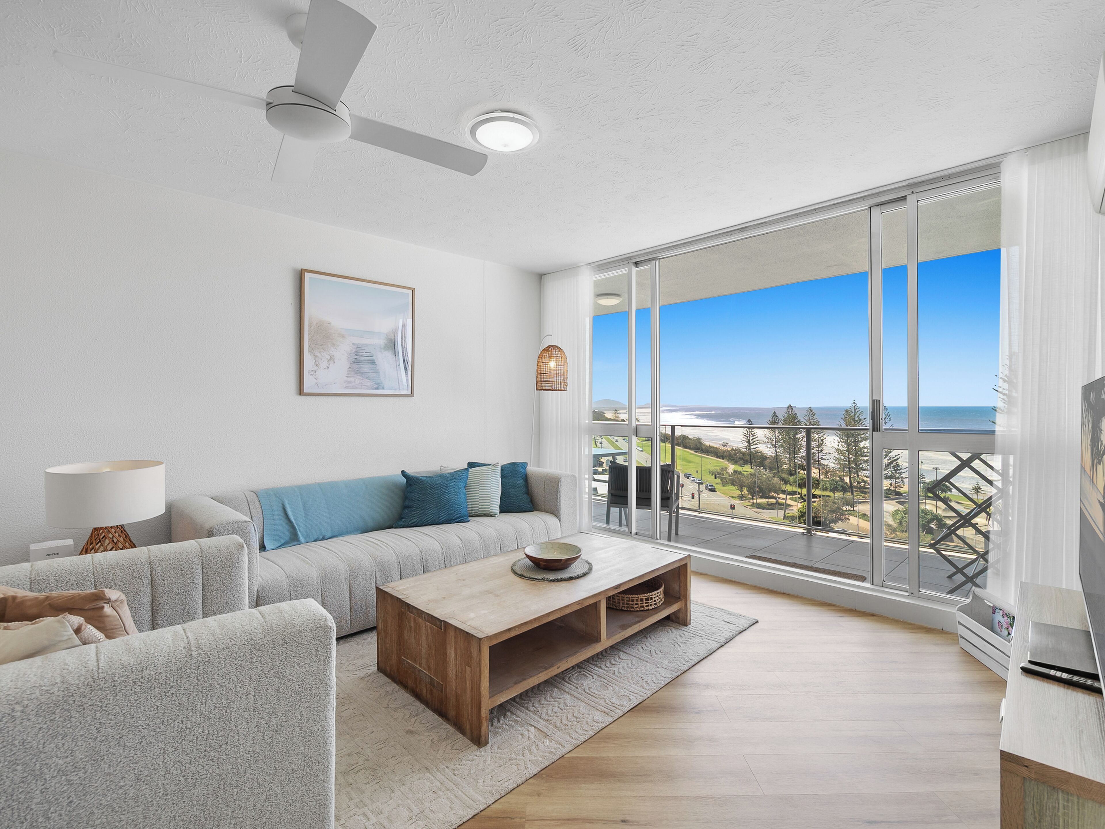 Uma sala de estar moderna com sofá, mesa de centro e uma janela grande com vista para a praia.