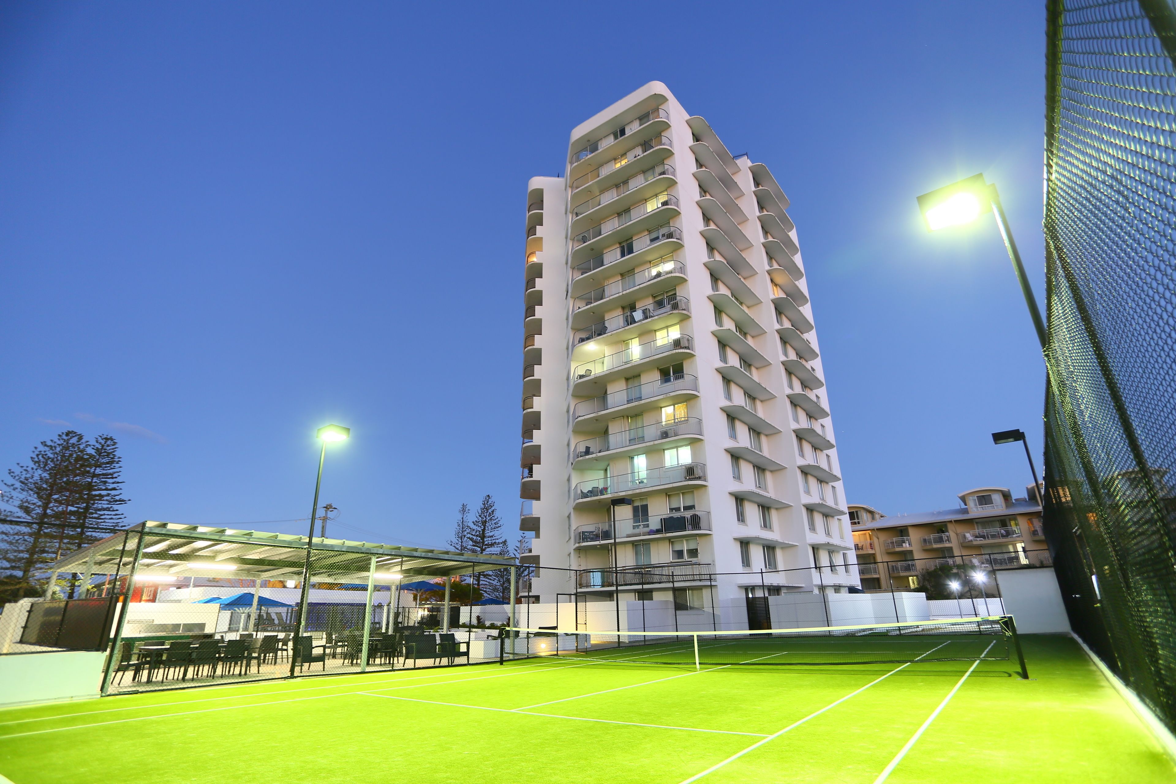 A tennis court with a net, surrounded by a fence, and a tall building in the background.