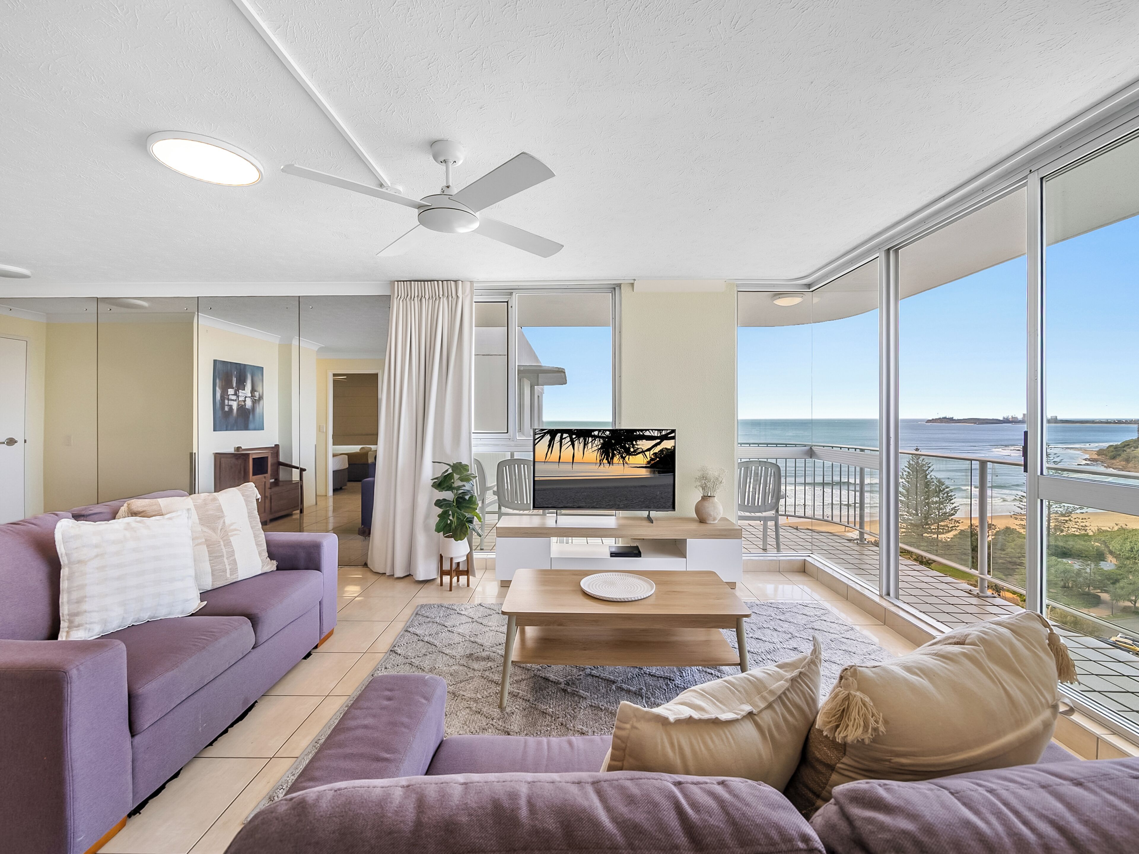 A modern living room with a purple sofa, a coffee table, and a TV. There is a balcony with a view of the ocean.
