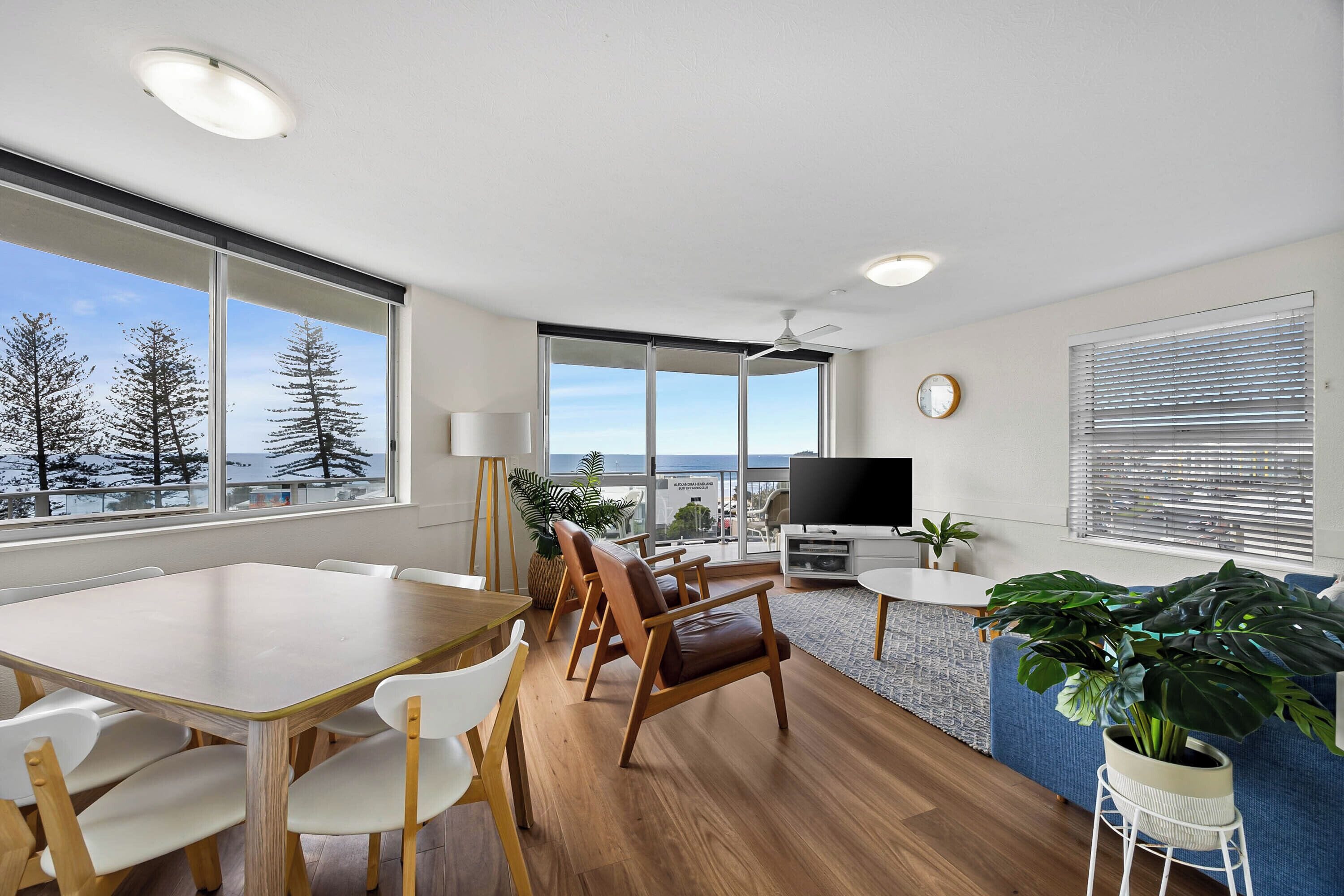Uma sala de estar moderna com piso de madeira, uma mesa de jantar com cadeiras, um sofá, uma televisão e janelas amplas que oferecem vista para árvores e o céu.