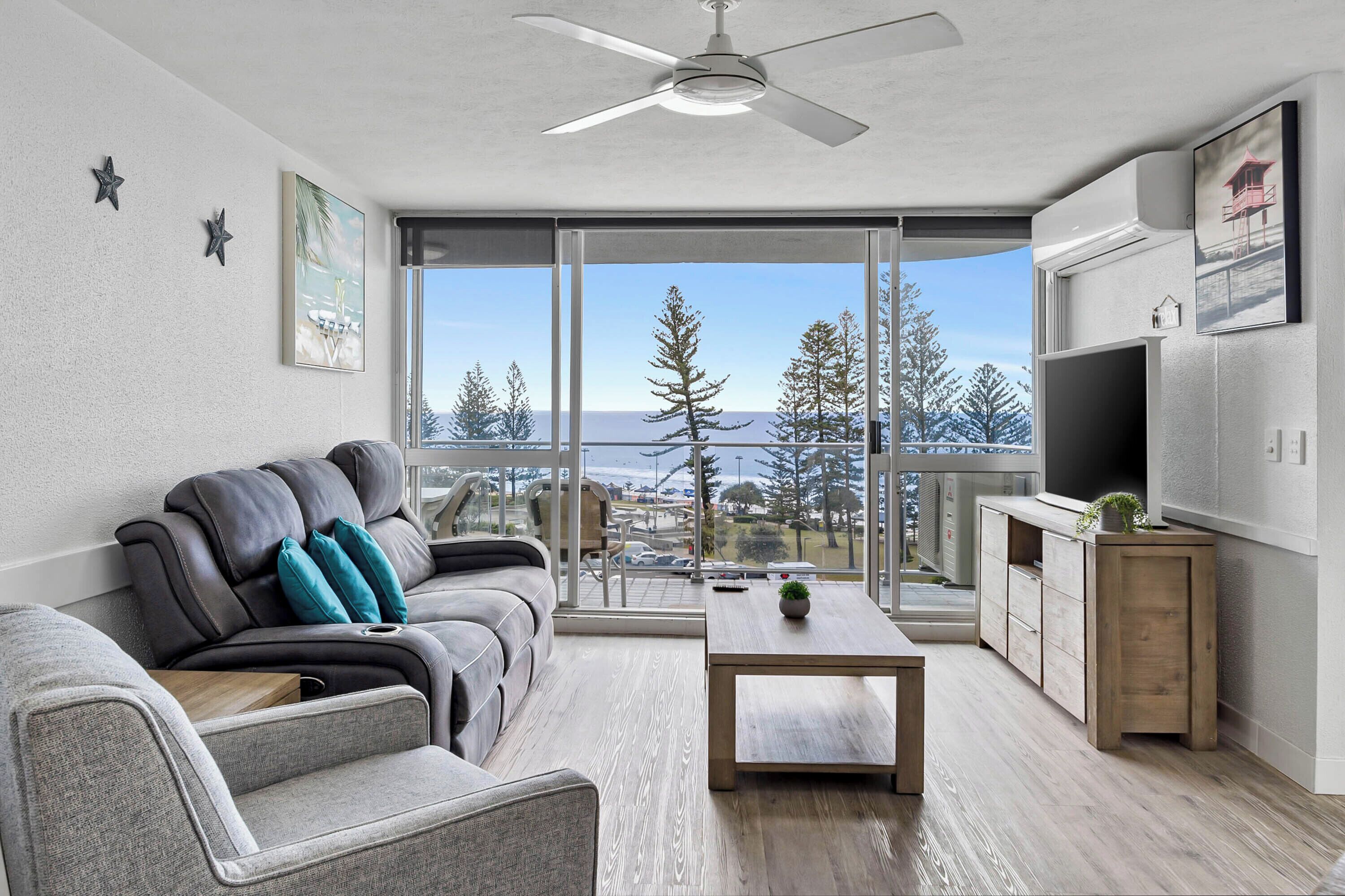 A modern living room with a sofa, a coffee table, and a TV. There is a balcony with a view of trees and a building.