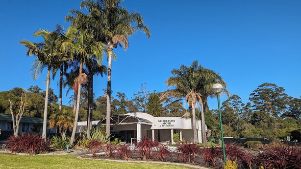 Gloucester Motel & Restaurant med et overdækket udendørs siddeområde, omgivet af palmer og anlagte haver.