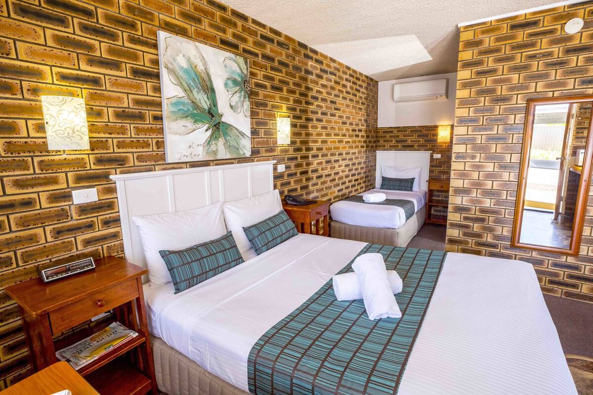 Une chambre d’hôtel avec un mur en briques, un grand lit avec une literie blanche, une table de chevet avec une radio et un miroir sur le mur.