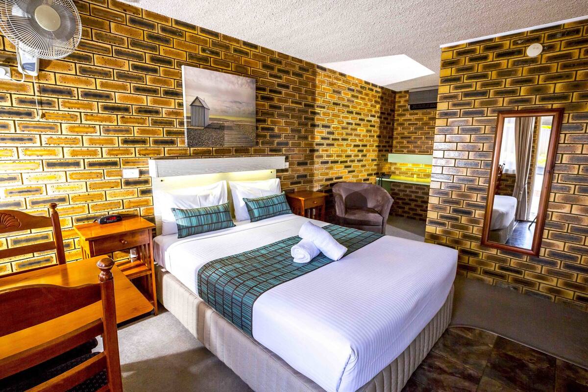Une chambre d’hôtel avec un mur décoré d’un motif en briques, un lit avec une literie blanche, une chaise en bois, un bureau et un miroir.