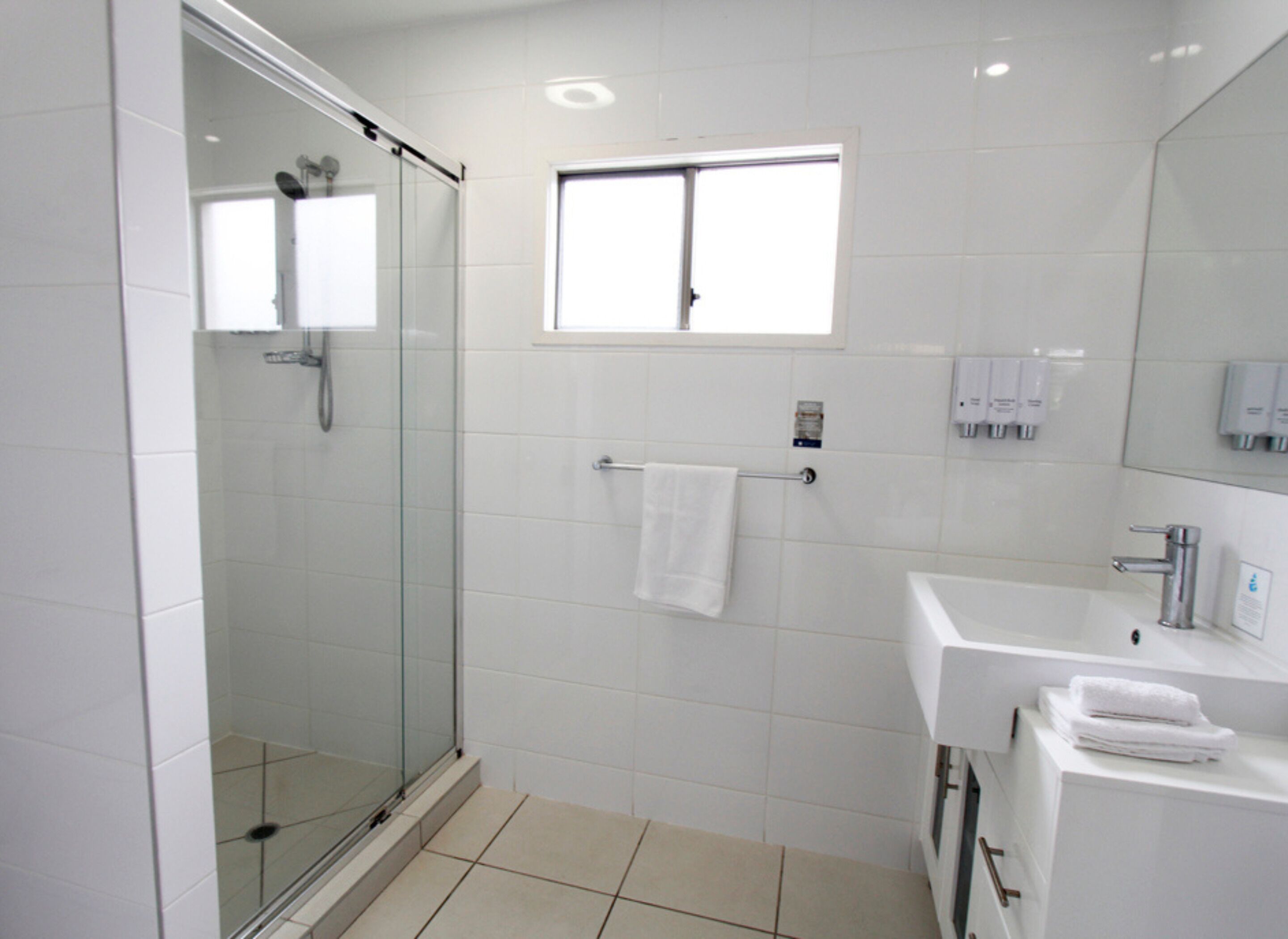 Ein modernes Badezimmer mit einer gläsernen Duschkabine, einem weißen Waschbecken mit Spiegel und einem Fenster darüber.