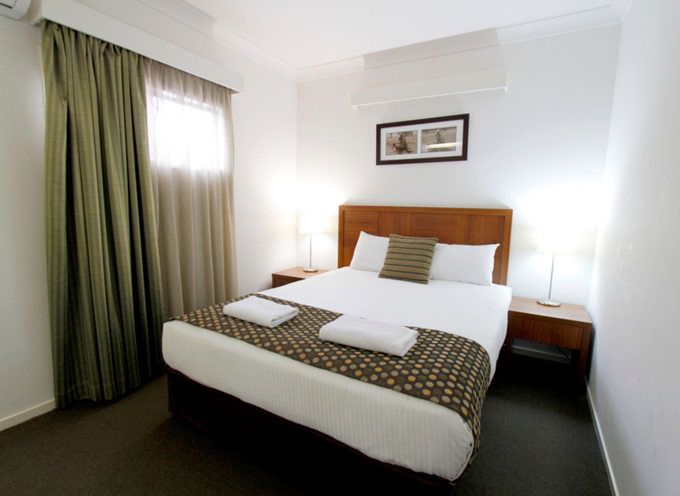 A hotel room with a large bed, two bedside tables, a headboard, a window with curtains, and a framed picture on the wall.