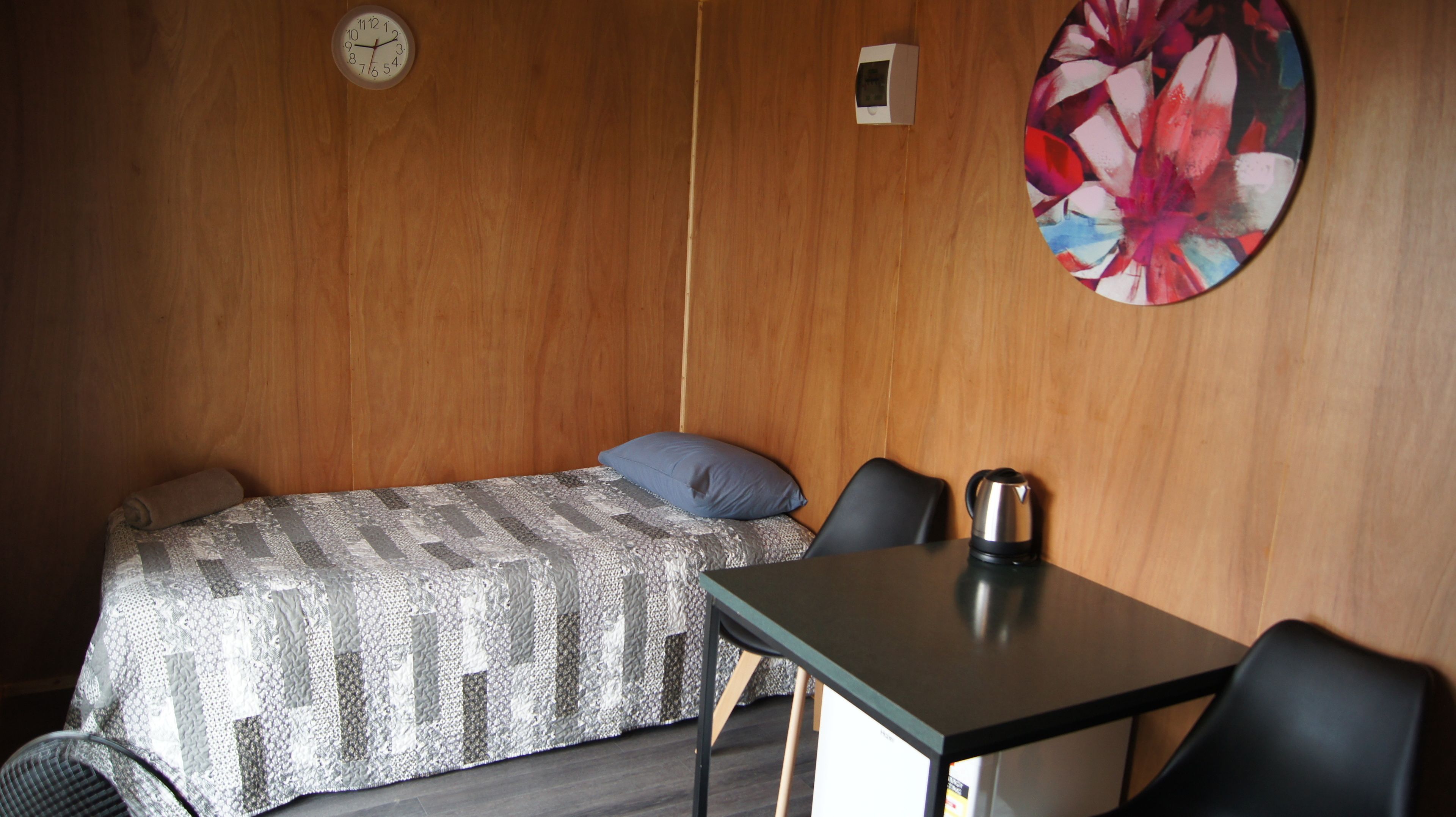 A small, neatly arranged room with a bed, a chair, a table, a kettle, and a clock on the wall.