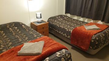 Ein Zimmer mit zwei Betten, jedes mit einer gemusterten Tagesdecke und einer Decke. Zwischen den Betten steht ein kleiner Nachttisch mit einer Lampe.