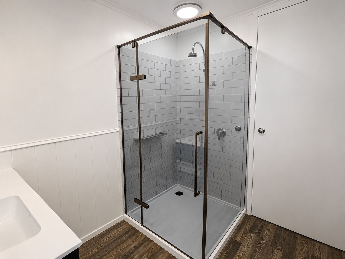 A glass-enclosed shower with a white tiled wall, a wooden floor, and a white bathtub.