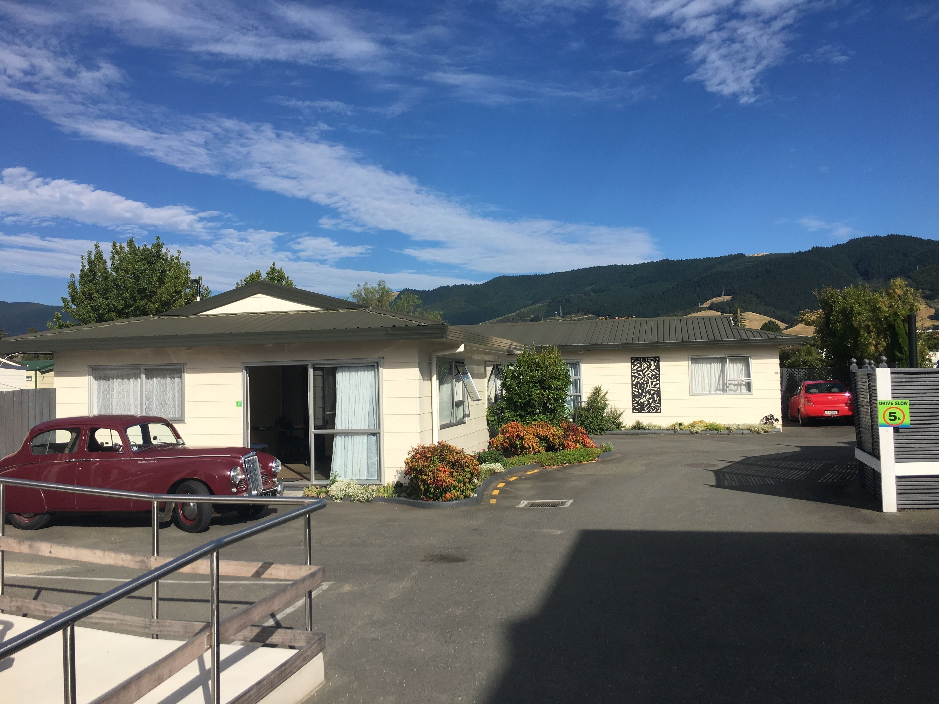 A motel with a red car parked in front, a small building with a door open, and a mountain in the background.