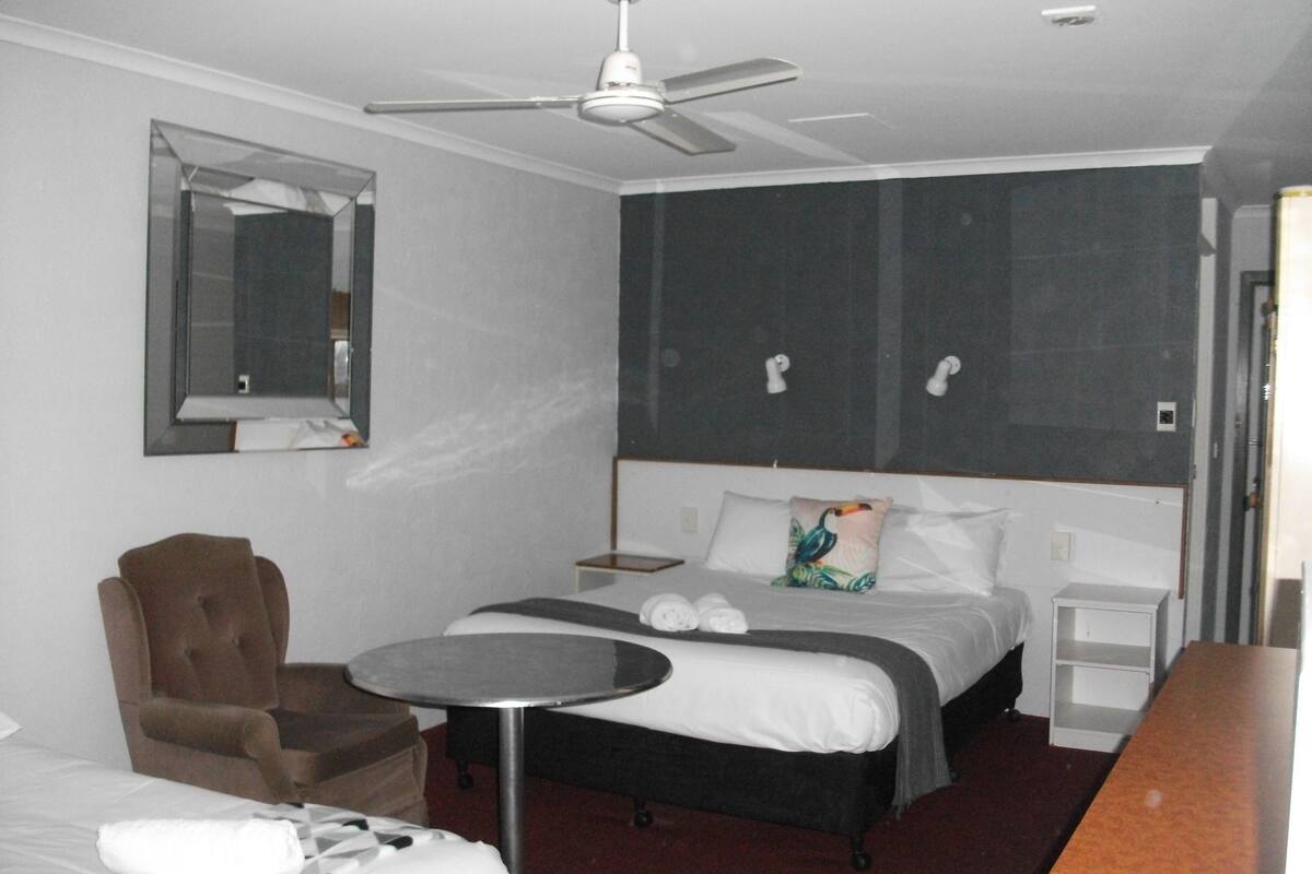 Habitació d'hotel amb un llit, una cadira, una taula rodona petita, un ventilador de sostre, un mirall i una tauleta de nit.