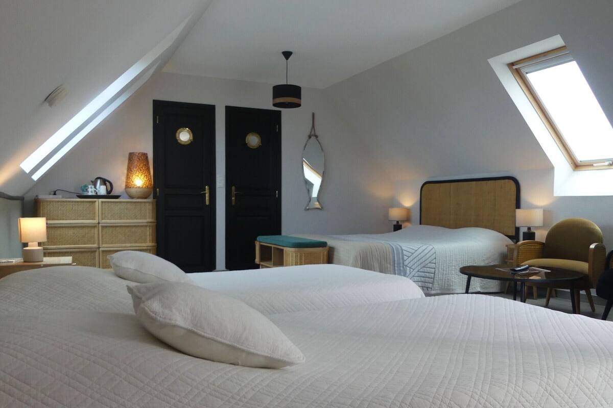 Une chambre avec deux lits, une petite table, une chaise et un bureau avec une lampe. Il y a deux portes et une fenêtre.