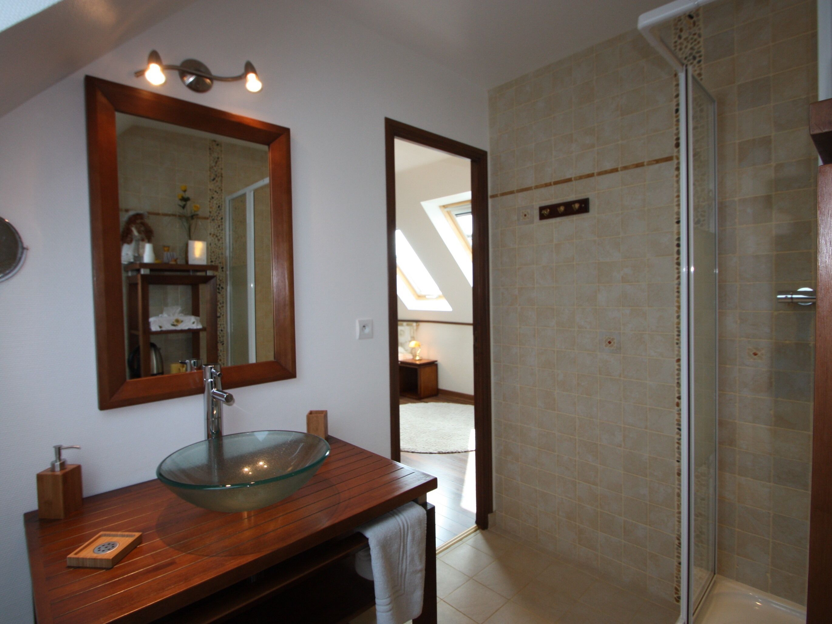 Un baño con lavamanos de vidrio, tocador de madera, un espejo grande y zona de ducha.