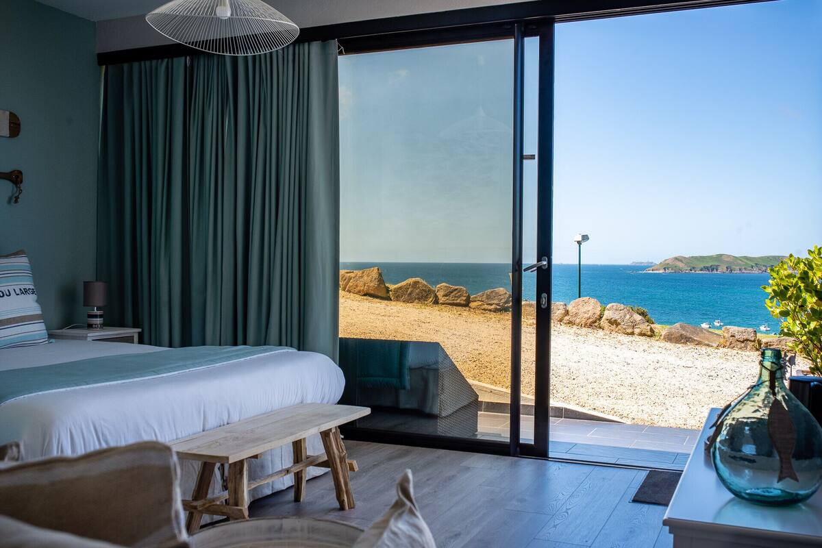 Une chambre dotée d’un grand lit, d’un banc en bois, d’une table de chevet et offrant une vue sur la plage et la mer grâce à une porte-fenêtre coulissante.
