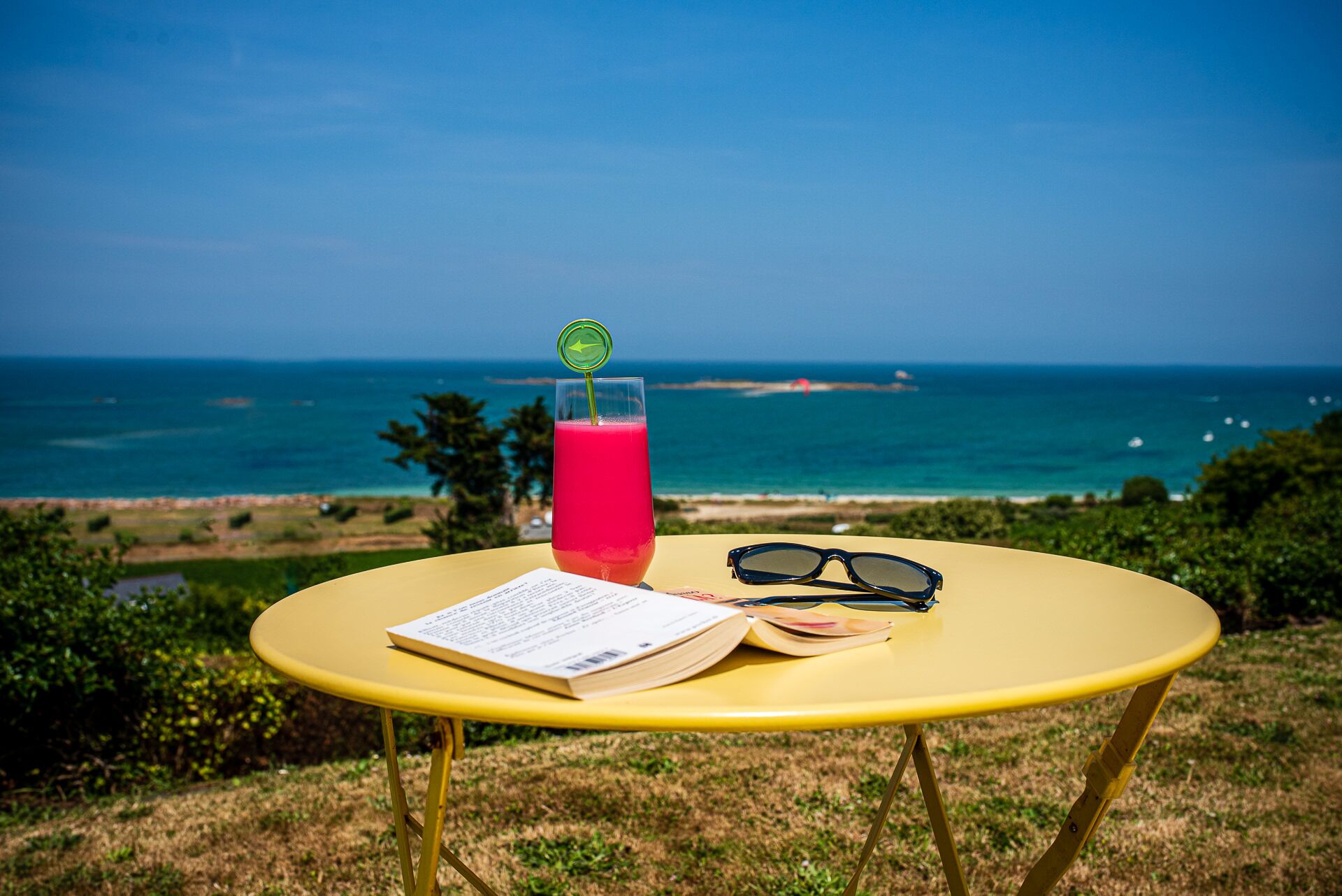 A yellow table with a drink, sunglasses, and a menu on a grassy hill overlooking a beach.