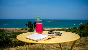 A yellow table with a drink, sunglasses, and a menu on a grassy hill overlooking a beach.
