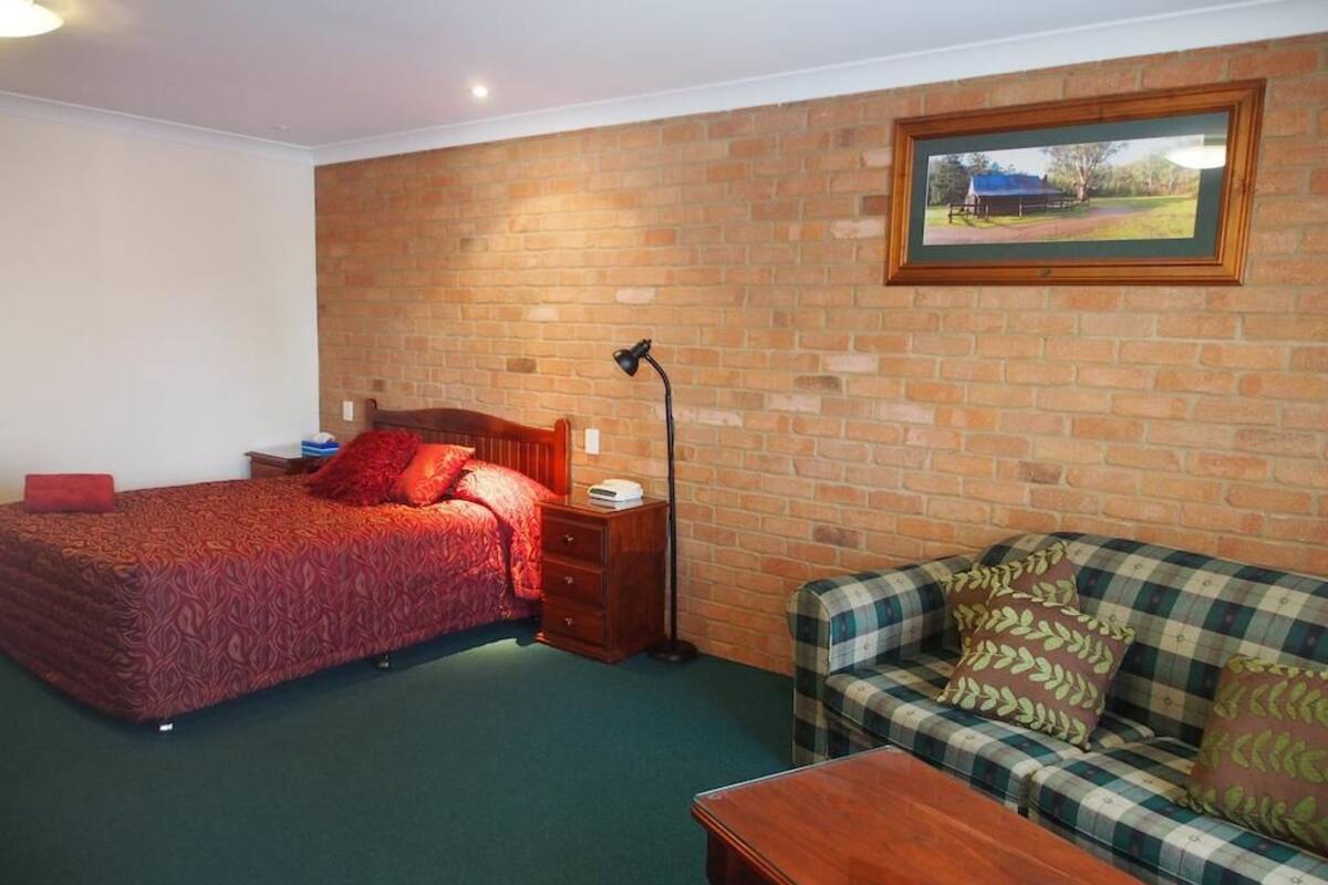 A room with a bed, a wooden nightstand, a lamp, a sofa, and a framed picture on the brick wall.