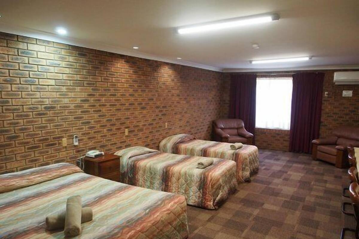 A hotel room with brick walls, three beds, a leather armchair, and a window with curtains.