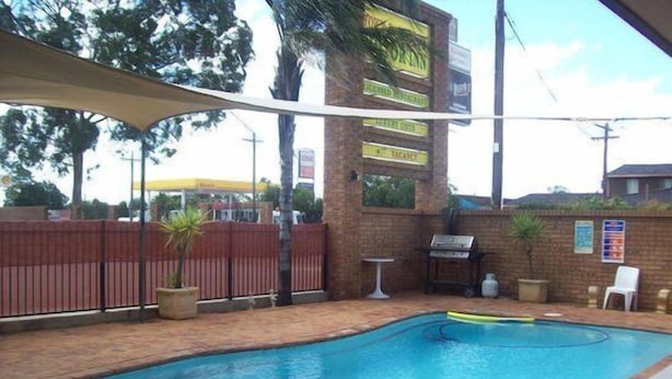 An outdoor swimming pool area with a brick patio, a barbecue grill, and a shaded seating area.