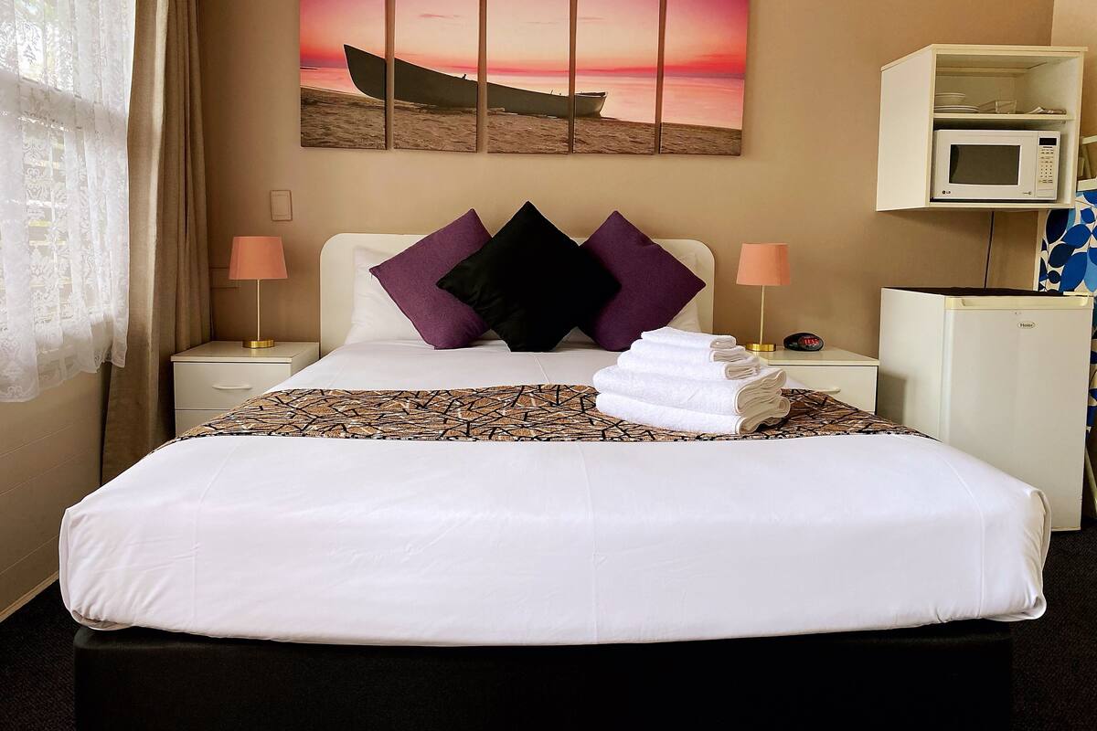 A hotel room with a bed, a painting of a boat on the beach, a microwave, and a small refrigerator.