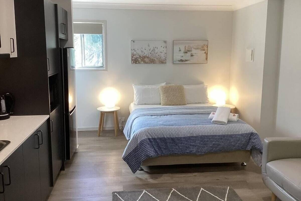 Un dormitorio compacto con cama, lámparas de noche, un pequeño taburete y una kitchenette con armarios y una cafetera.