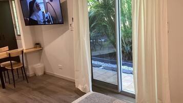 A room with a bed, a TV mounted on the wall, a small table with chairs, and a sliding glass door leading to an outdoor area with plants.