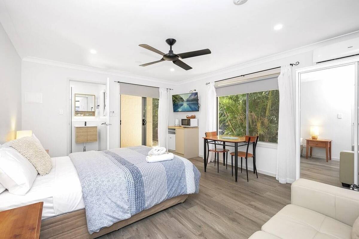 Un dormitorio moderno con cama, ventilador de techo, comedor con mesa y sillas, y vista al exterior.