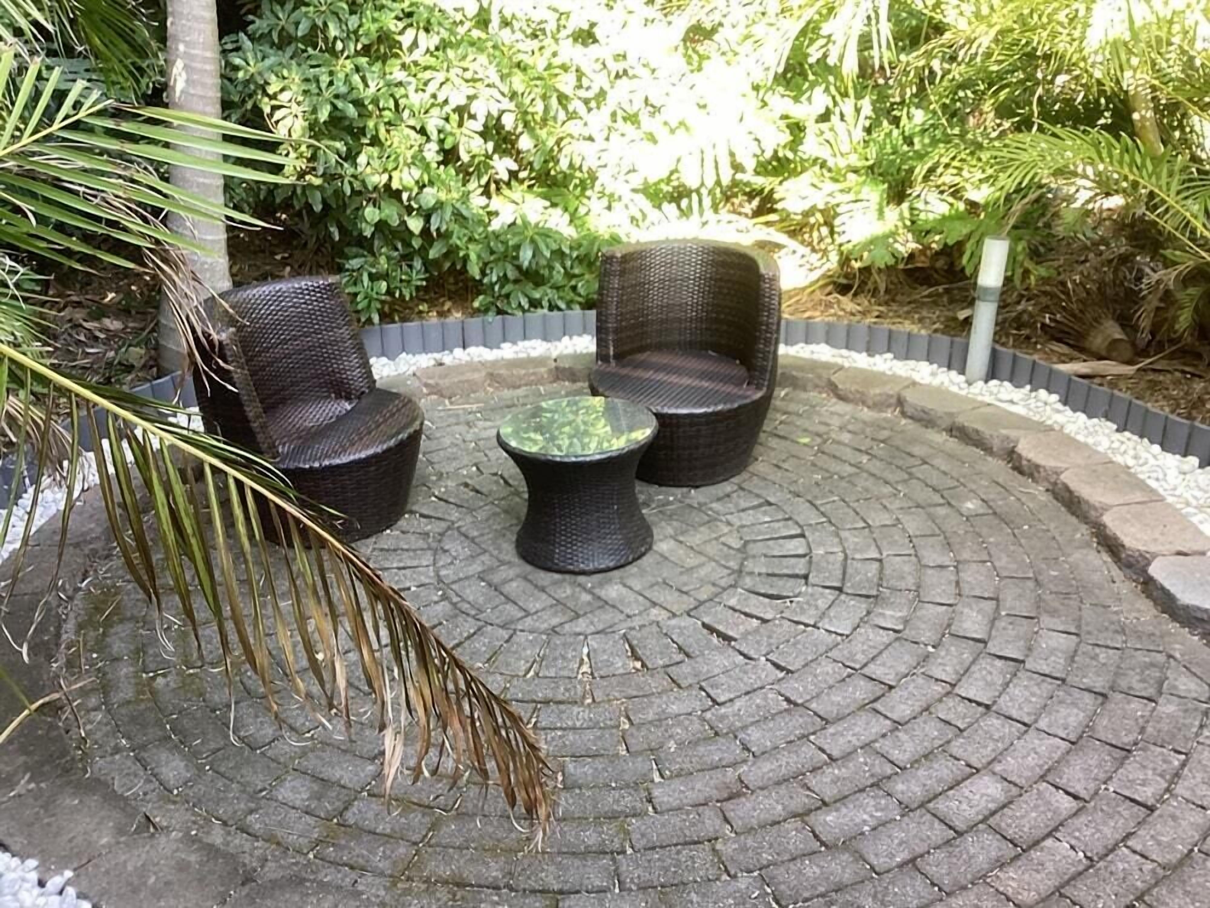 A circular outdoor seating area with wicker furniture and a glass table, surrounded by a brick border and greenery.