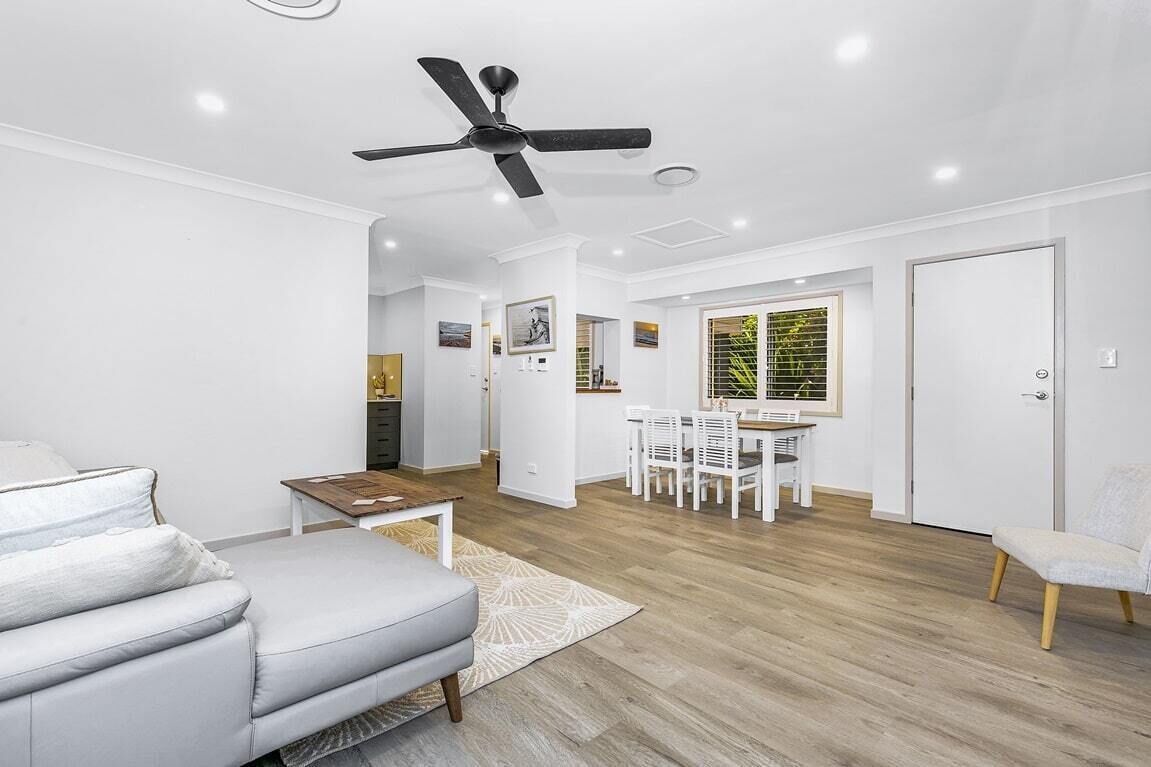 A modern living room with a sofa, a dining table with chairs, a ceiling fan, and a view of the outside through a window.