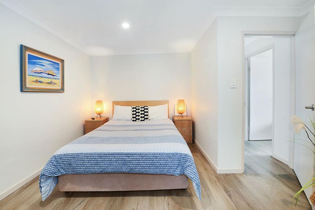 Un dormitorio ordenado con una cama, dos mesitas de noche con lámparas, un cuadro en la pared y una puerta que da a otra habitación.