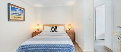 Une chambre bien rangée, avec un lit, deux tables de chevet dotées de lampes, un tableau encadré au mur et une porte donnant sur une autre pièce.
