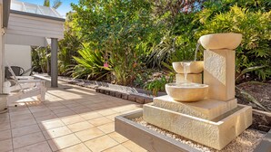 A patio area with a stone fountain, a white outdoor seating area, and lush greenery. - Tiarri Terrigal (Terrigal)