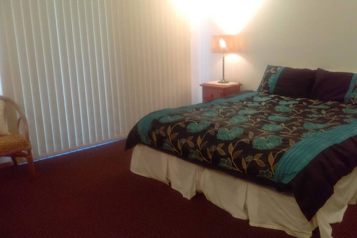 A bedroom with a bed, a nightstand, a lamp, a chair, and a window with blinds.