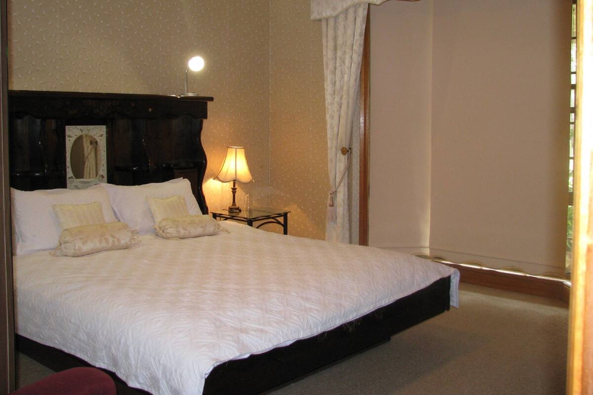 A neatly made bed with a white comforter and pillows, a bedside table with a lamp, a mirror, and a window with curtains.