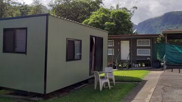 Una piccola casetta a un piano con una porta e finestre, un tavolino con sedie, e un'area coperta da un telo verde.