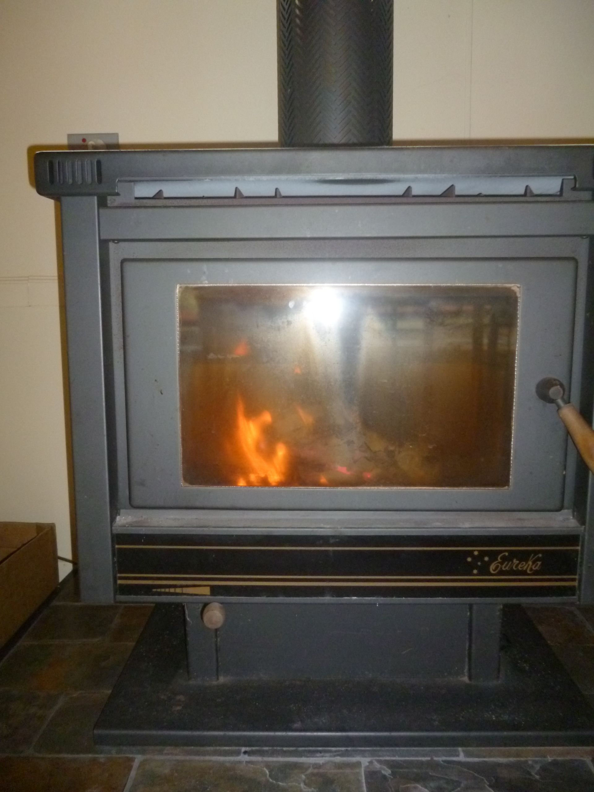 Una estufa de leña con puerta de vidrio, una chimenea de metal y un asa en el lado derecho.