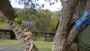 Un arbre orné de sculptures de grenouilles et de papillons, un bâtiment avec un toit rouge et des fleurs violettes qui poussent sur le sol.