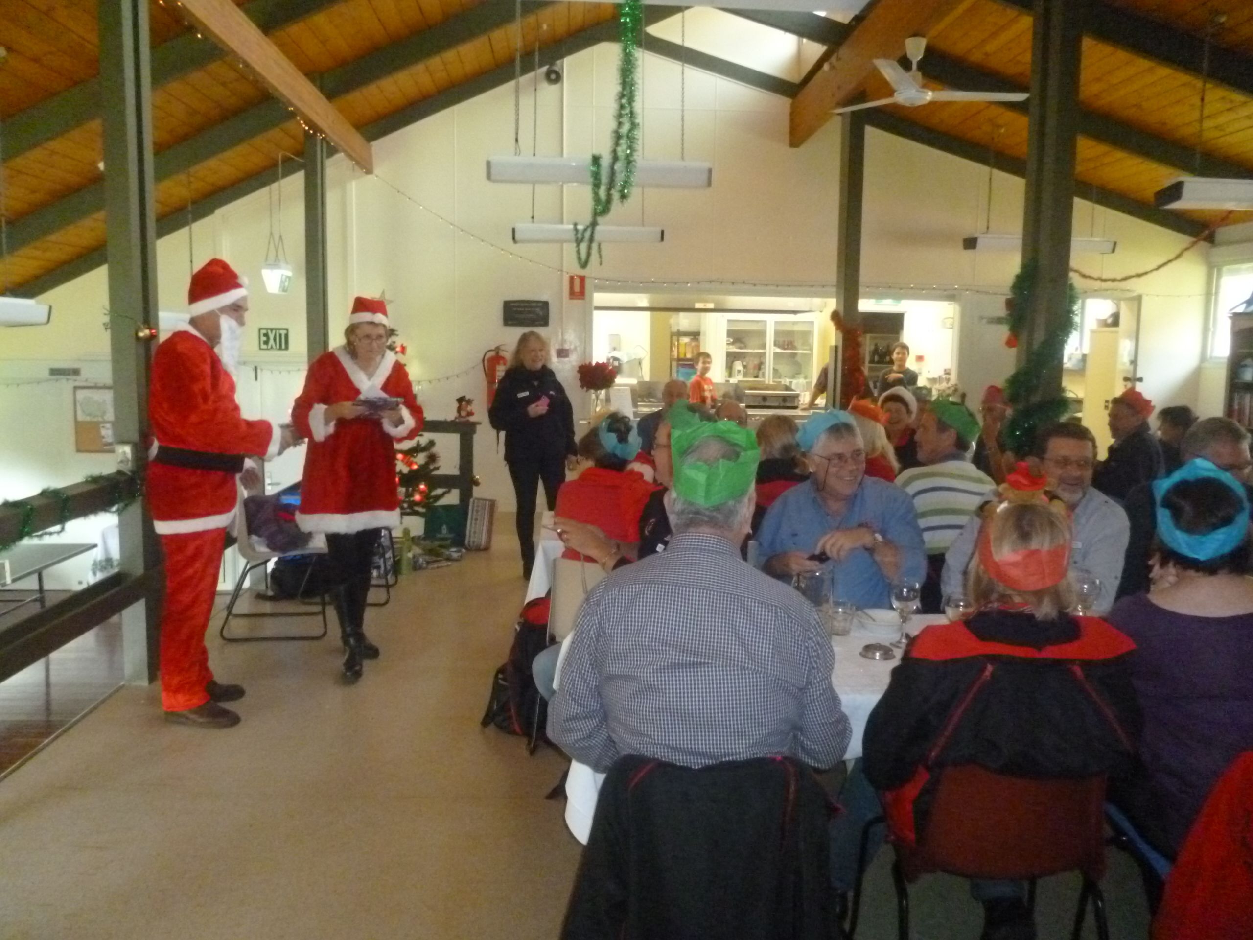 A festive indoor gathering with people seated at tables, a person in a Santa costume, and Christmas decorations.