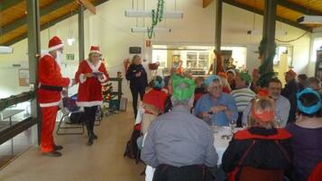 A festive indoor gathering with people seated at tables, a person in a Santa costume, and Christmas decorations.