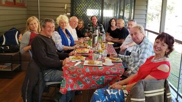 Un grupo de personas sentadas alrededor de una mesa con mantel rojo, suelo de madera y una ventana que da a zonas verdes.