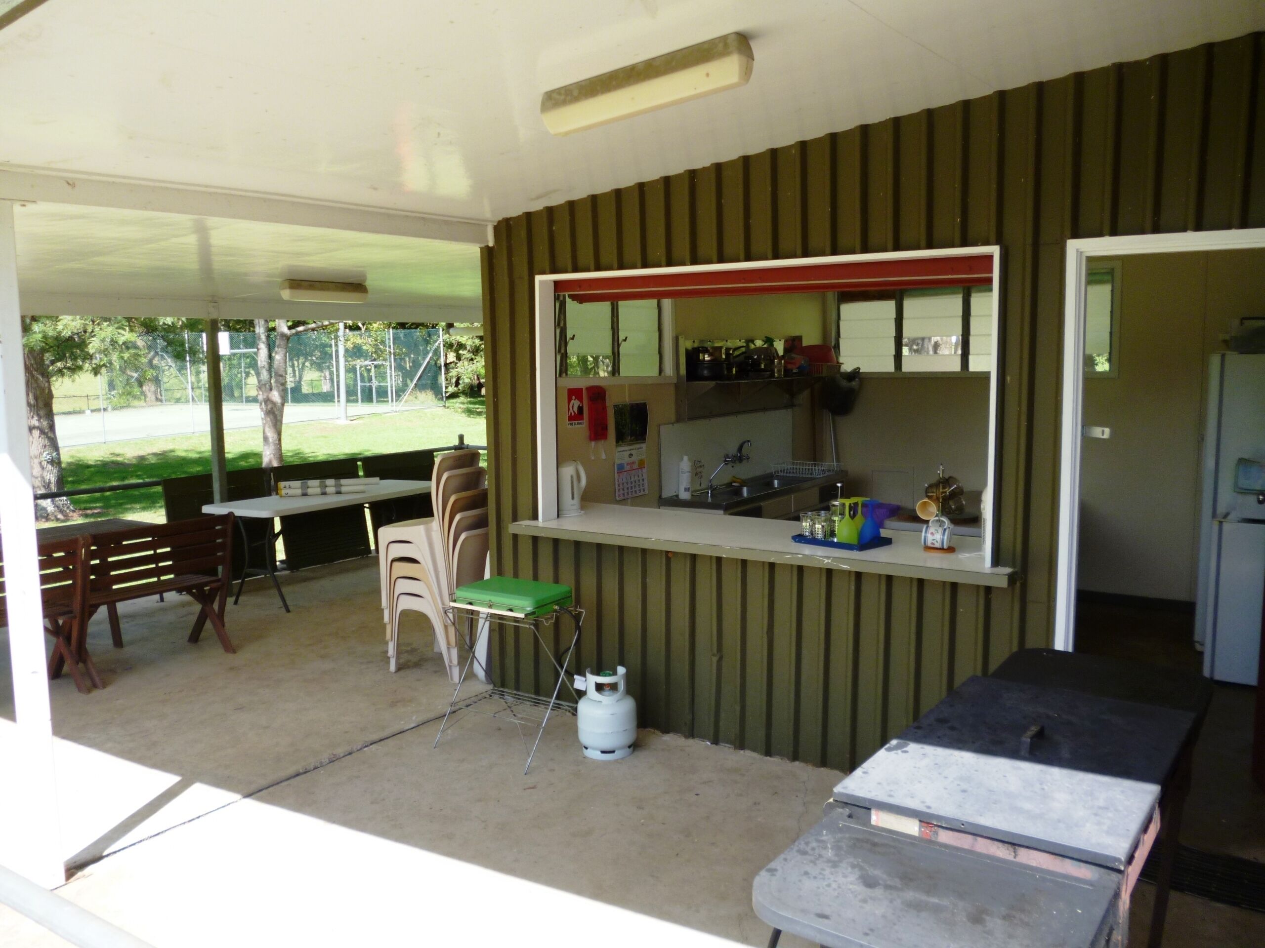 A covered outdoor area with tables and chairs, a gas cylinder, and a refrigerator.