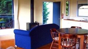 Una habitación con un sofá azul estampado, una mesa de comedor de madera con sillas, una zona de cocina con estufa y ventanas amplias que dan a zonas verdes.