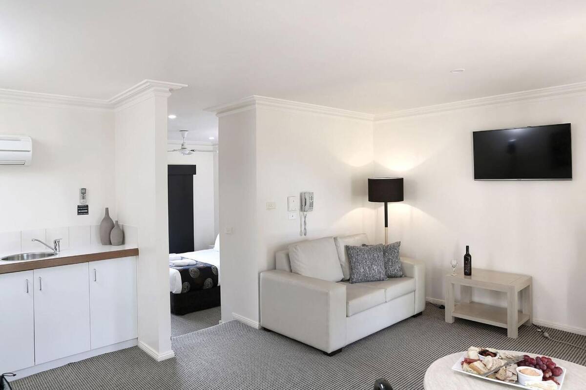 A modern hotel room with a kitchenette, a sofa, a TV, and a small table with a bottle and glass.