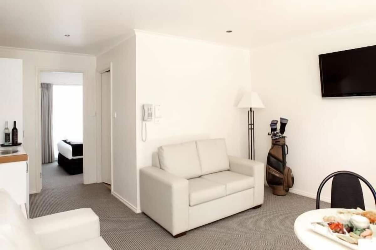 A hotel room with a white sofa, a dining table with food, a lamp, a guitar, and a TV on the wall.