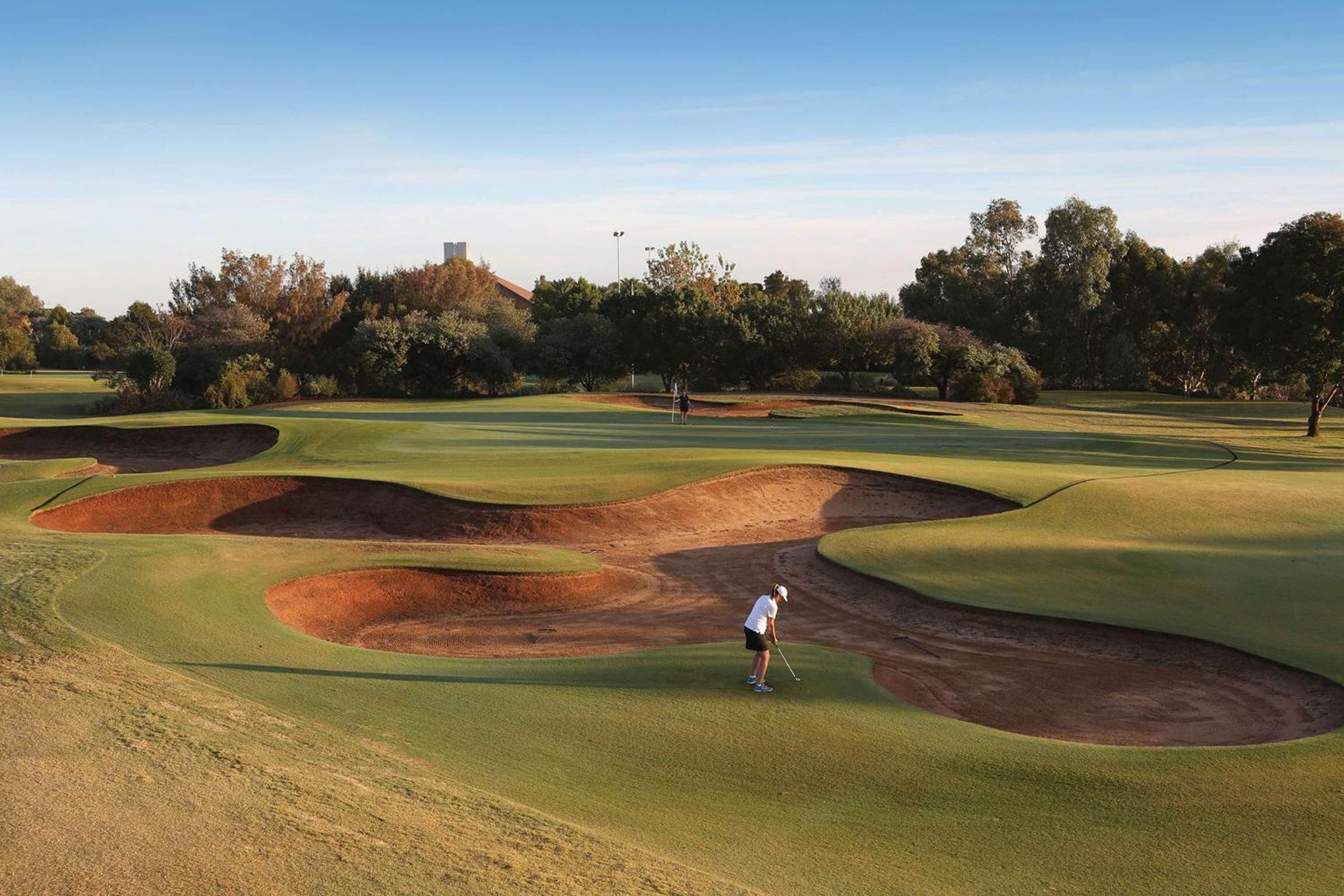 Um campo de golfe com uma pessoa prestes a dar tacada, rodeado por bunkers de areia e árvores.