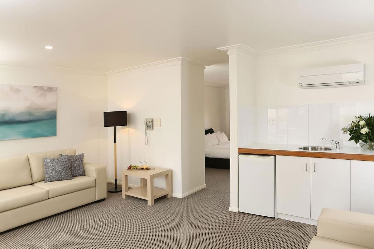 A modern hotel room with a living area, a small kitchen, a bed, and a painting on the wall.