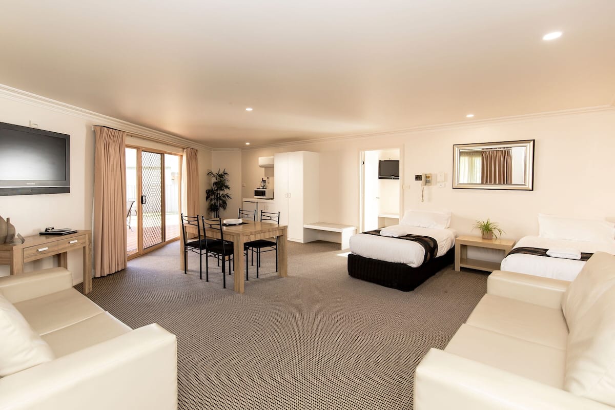 A hotel room with a bed, two white sofas, a dining table with chairs, a TV mounted on the wall, and a large mirror.