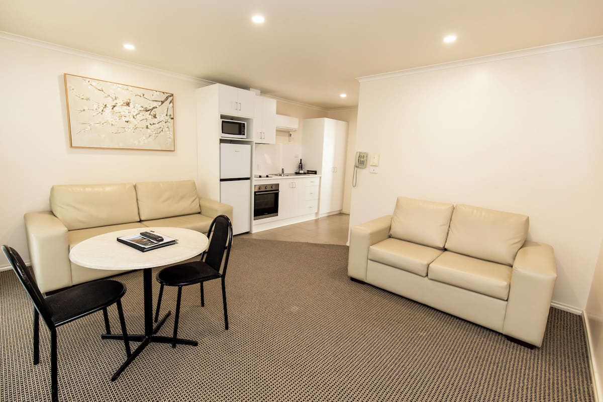 A modern living room with a beige sofa, a round coffee table, and a black chair. There is a kitchen area in the background with white cabinets and appliances.