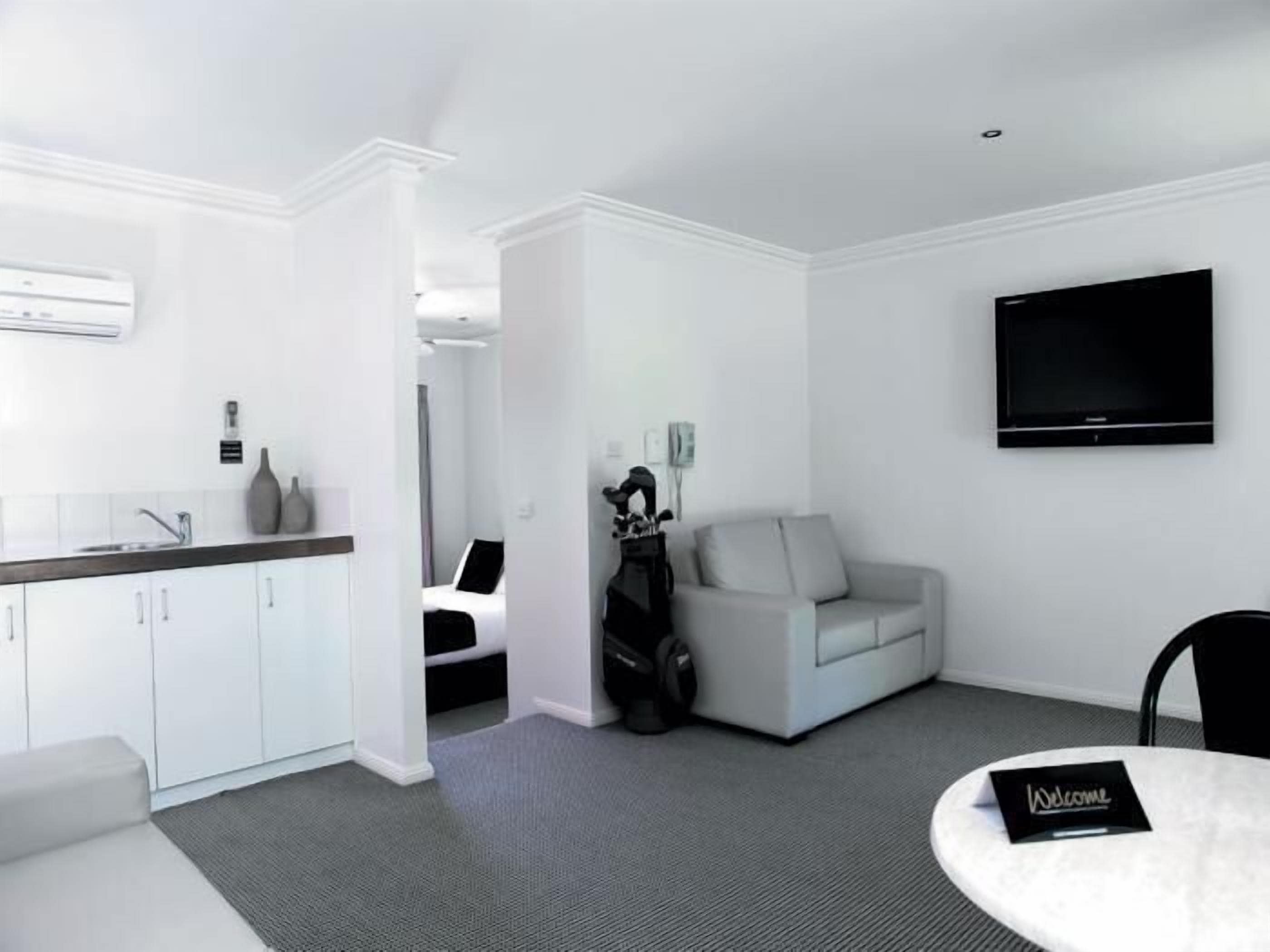 A modern hotel room with a kitchenette, a flat-screen TV, a sofa, and a small table with a 'Welcome' sign.