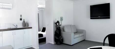 A modern hotel room with a kitchenette, a flat-screen TV, a sofa, and a small table with a 'Welcome' sign.